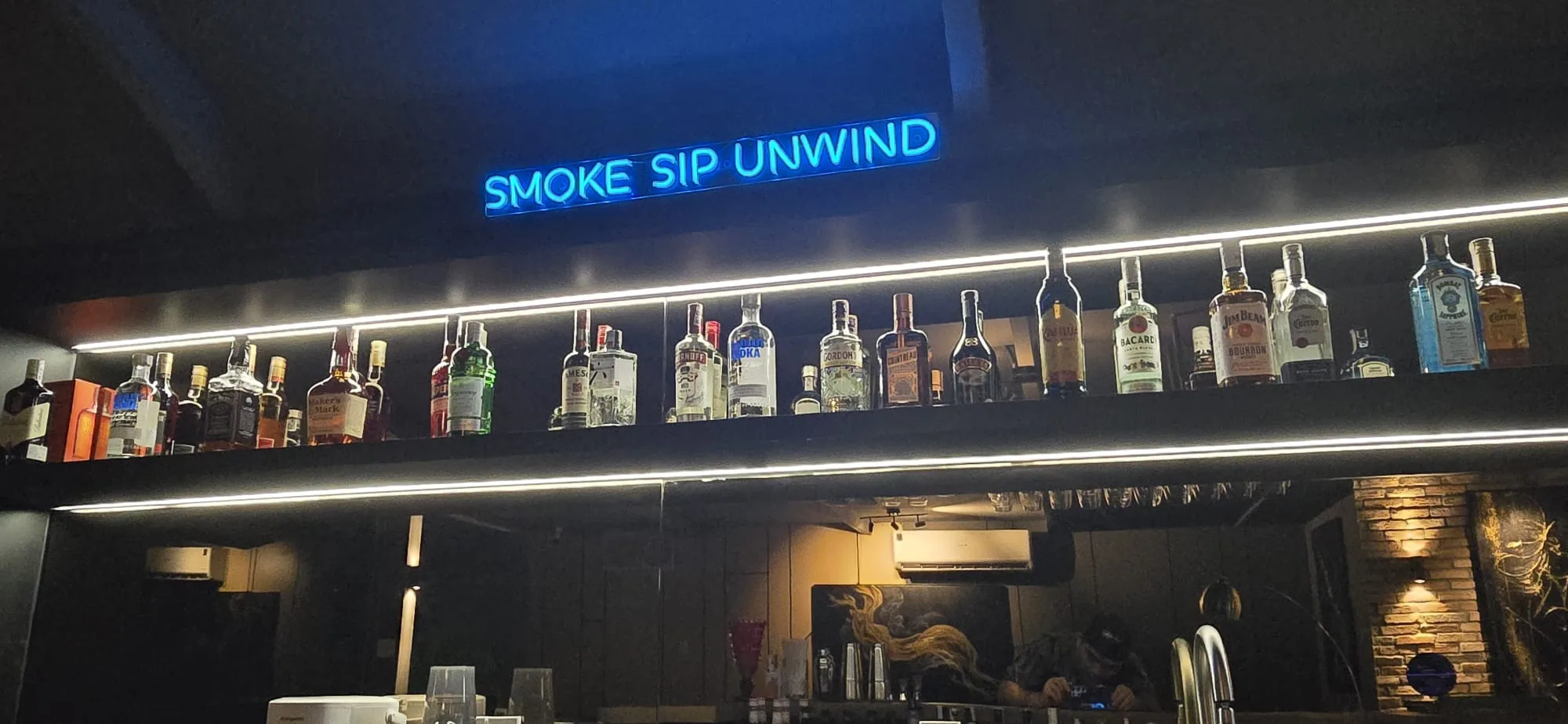 Smoke Sip Unwind neon sign above the backlit bottle shelf at Natasha Noir